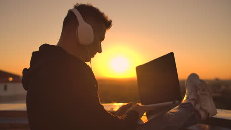 A-man-freelancer-in-headphones-standing-on-the-roof-at-sunset-writes-on-the-keyboard-code-pages.-Little-business.-Listen-to-music-and-work-at-the-computer-enjoying-the-beautiful-view-from-the-roof.